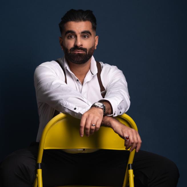 Tez Ilyas dressed in a white shirt facing the camera sat backwards on a yellow chair.