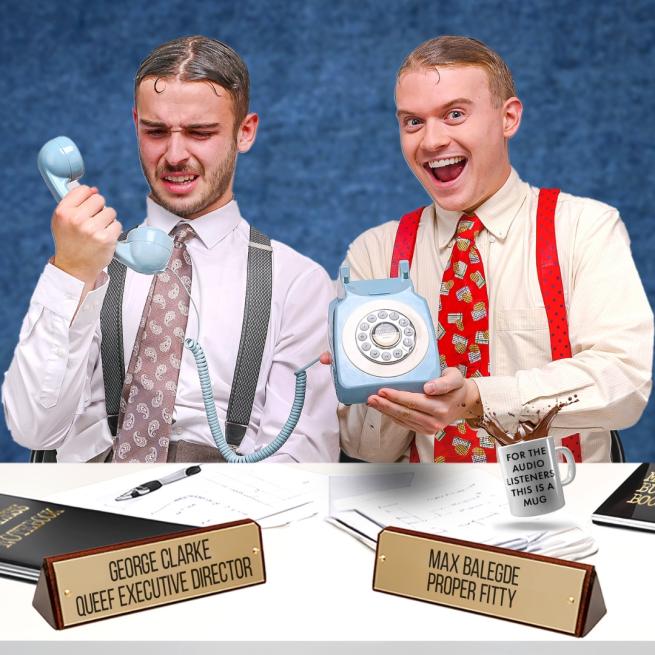 Two men in shirts, ties and braces sat at a desk and holding up an old-fashioned dial-up phone. One looks confused and the other smiles widely. They have name plaques in front of the saying George Clarke Queef Executive Director and Max Baledge Proper Fitty