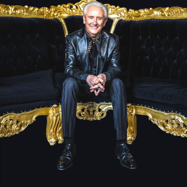 Tony Christie in a black leather suit jacket and black and gold neck tie sat on a black and gold sofa. 