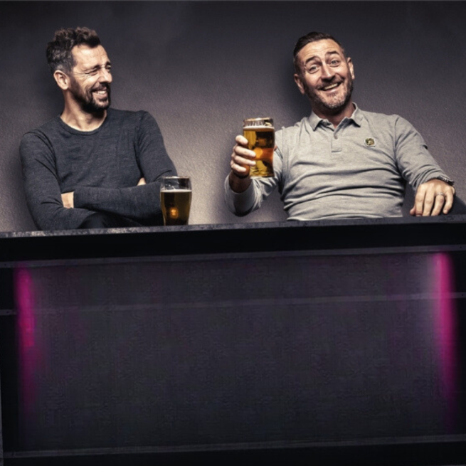Will and Ralf sitting at a bar counter both with pints and laughing with a plain grey background.