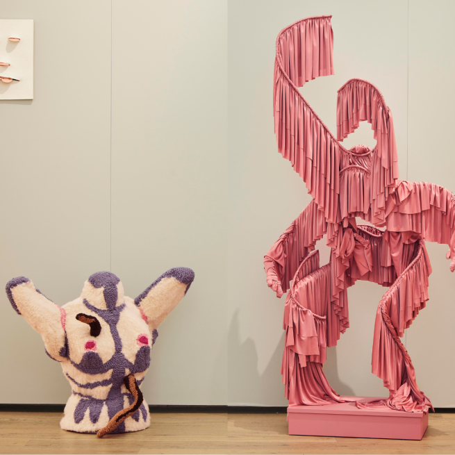 Two fabric sculptures against a white wall. On the left is a furry female torso and on the right is a figure made from wire and pink fabric fringing.