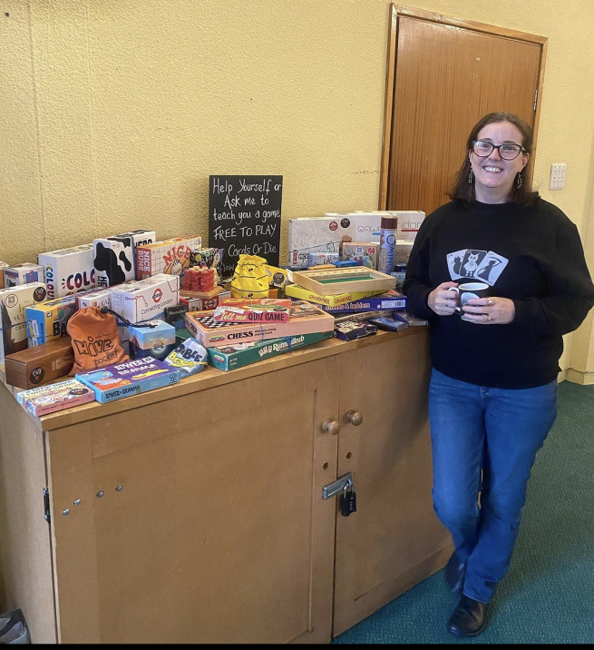 Me (a white woman with mid length dark hair, wearing glasses, a black jumper with the Cards or Die logo on and blue jeans. I am holding a cup of tea.) standing next to a selection of board and card games arranged on top of a large cupboard. 