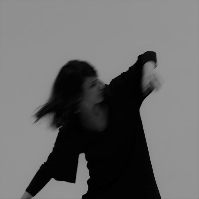 Black and white image of a woman with dark hair. Wearing all black, her arms are poised in movement and the image is slightly blurry to show her moving quickly