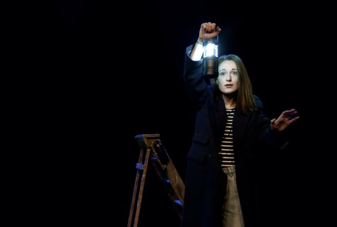 A woman holding up a lantern in the darkness and looking trepidatious 