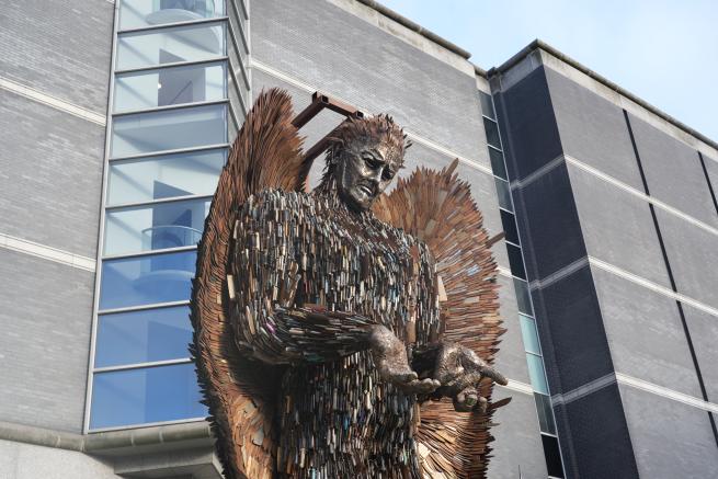 Metalwork sculpture made of knives in the shape of an angel with arms open and palms facing forwards in front of the Royal Armouries museum building