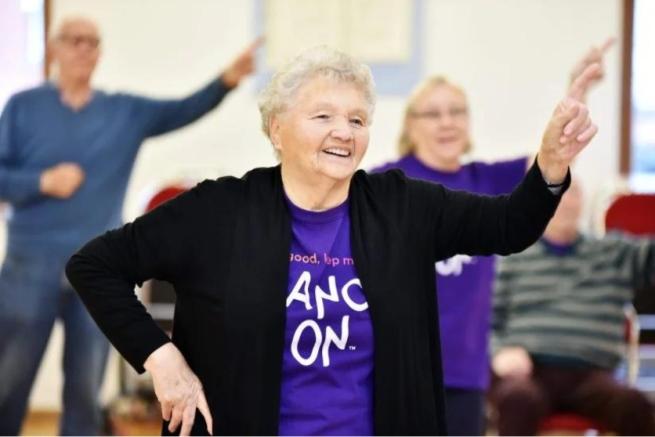 A white lady with short light hair strikes a disco pose with a big smile on her face. She wears a purple 'Dance On' Tshirt.