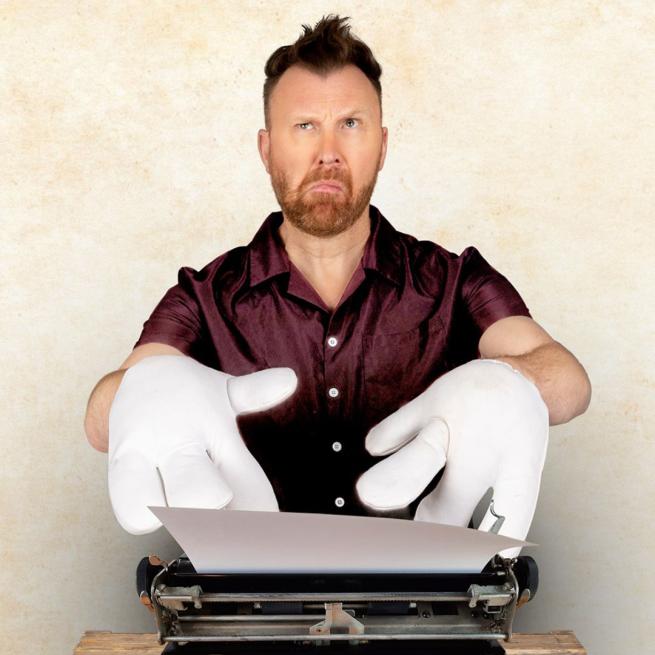 Jason Byrne wearing chunky white gloves typing on a typewriter looking confused.