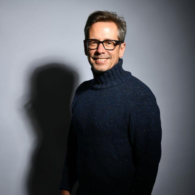 Nick Heyward smiling at the camera wearing a dark blue turtle neck jumper in front of a grey background.