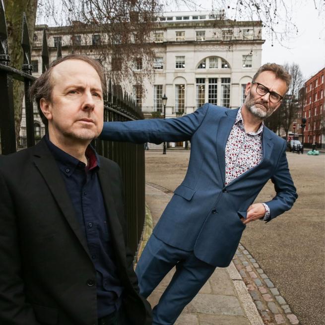 Steve Punt and Hugh Dennis on a street corner. Steve is leaning against a railing and Hugh is leaning off it behind him.