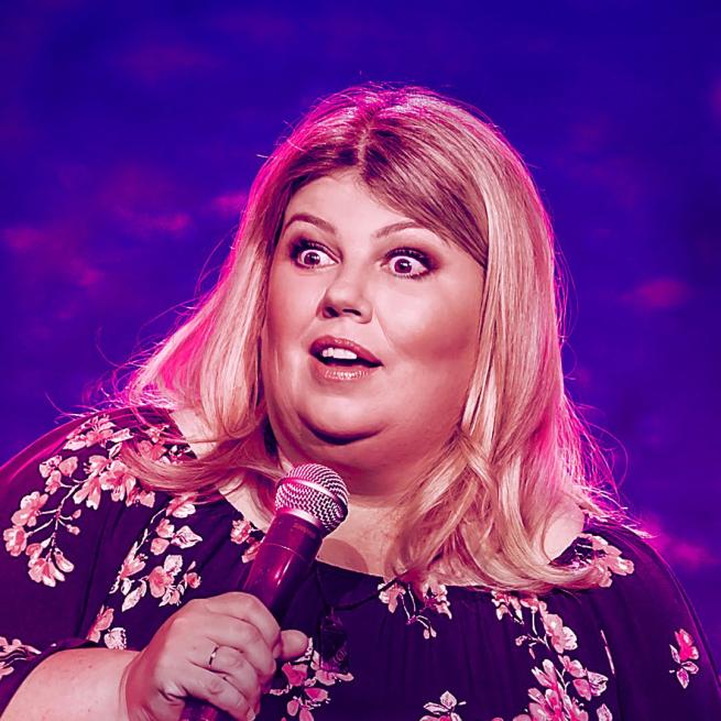 Urzila Carlson on stage looking shocked whilst holding a microphone. Pink-purple background.