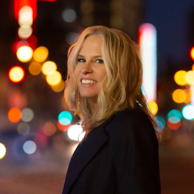 Vonda Shepard smiling, she is outside at night with glowing street lights behind her