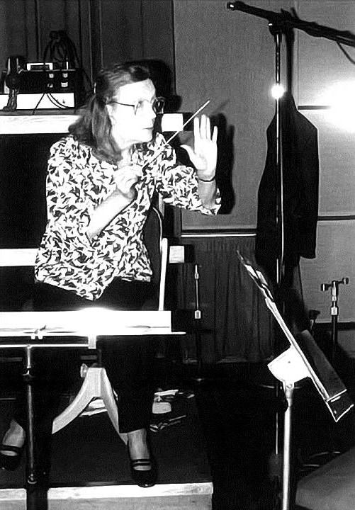 Anglea Morley conducting an orchestra. She is white with long dark hair, large glasses and a patterned shirt. She is waving a conductors baton.
