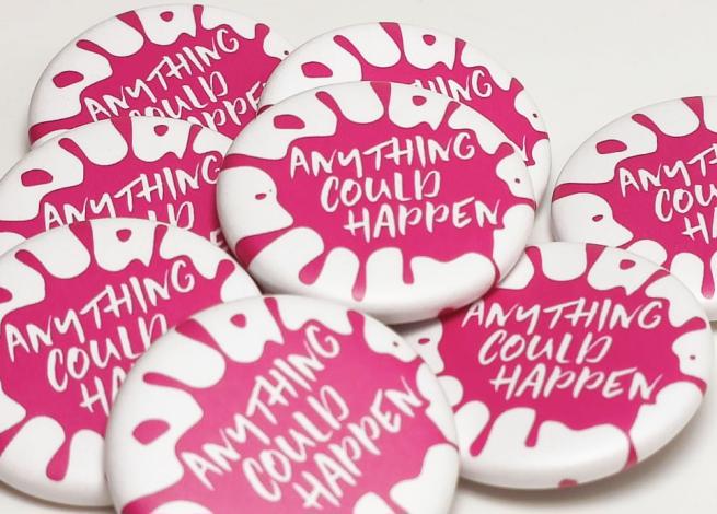 A close up of a pile of badges with an Anything Could Happen pink logo 