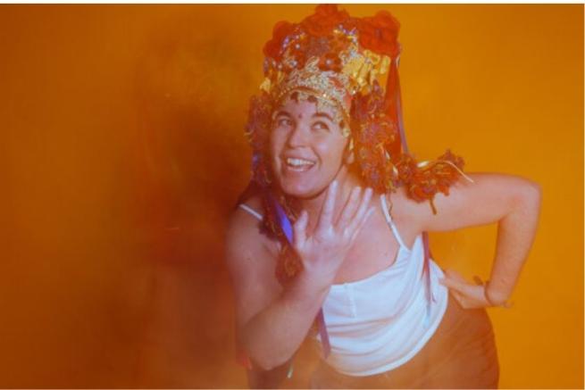 person in a beaded headdress and white tank top dancing against an orange backdrop