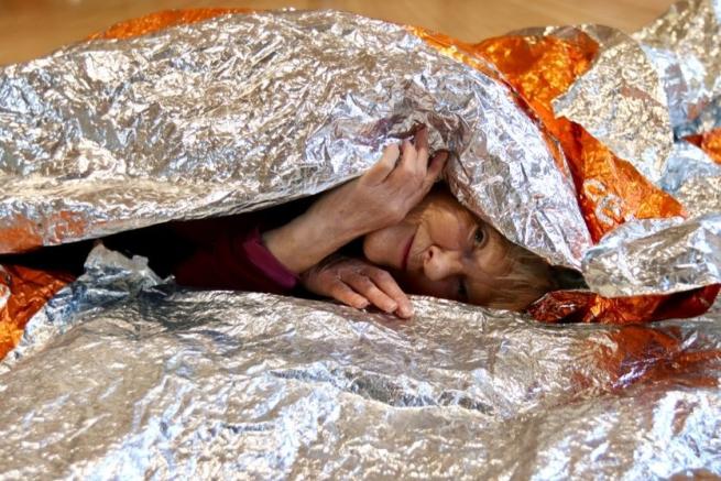 Elderly woman peeking through an orange and silver foil blanket, lied down and smiling.