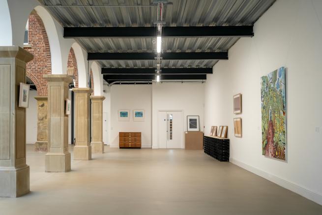 A section of the gallery space with arches and stone columns on the left and a white wall in the far end and to the right. There are artworks hung on the walls, and propped on furniture. 