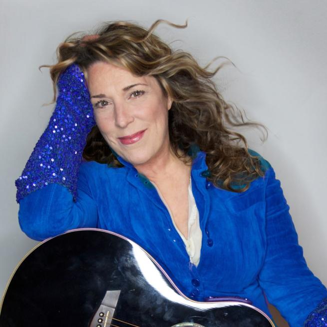 Beth Nielsen Chapman in a blue blouse and a black guitar with her right hand on the right side of her face.