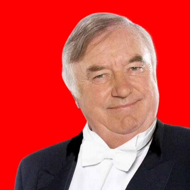 Jimmy Tarbuck from the chest upwards wearing a black suit and white bow tie on a red background.