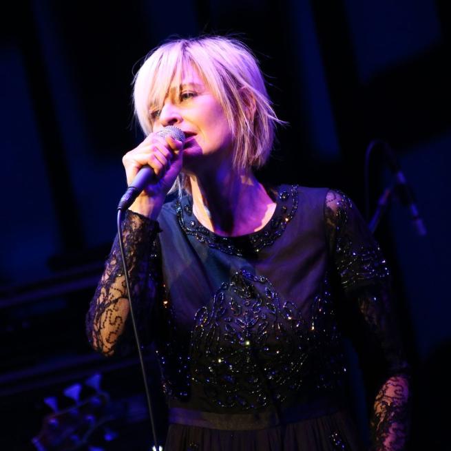 Julia Fordham on stage singing into a microphone in a black dress with a black sequin pattern on it.