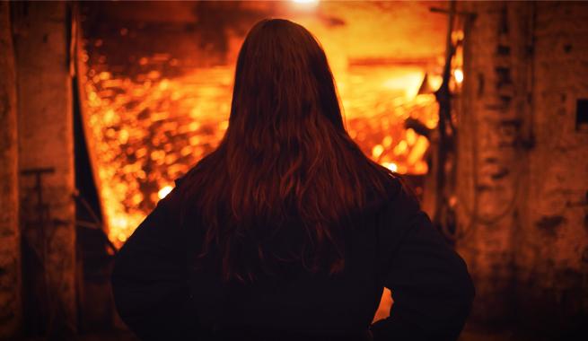 The back of a woman is in the foreground, turned to look at a forge, glowing hot with flame in the background