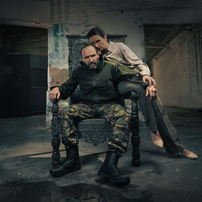 Ralph Fiennes as Macbeth sat on a metal throne wearing army camouflage trousers, dark green jacket and combat boots and Indira Varma as Lady Macbeth in a pale pink silk blouse, grey trousers and pale pink heels sat on the left arm of the chair. They are in an abandoned warehouse.