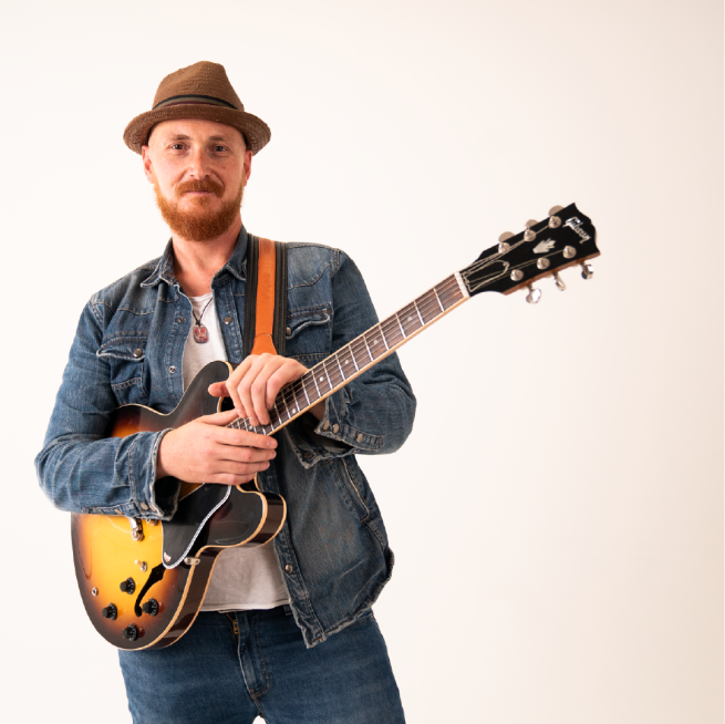 Paolo Fuschi wearing a denim jacket and trilby hat holds a guitar whilst smiling at the camera
