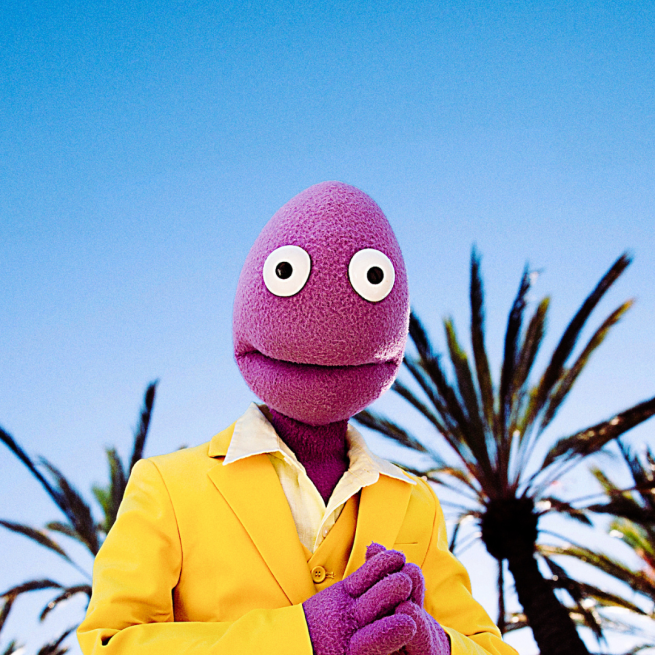 Randy Feltface pictured from the chest up wearing a bright yellow suit and his purple hands to his chest. His purple felt face and round eyes are staring right out to the camera in the centre of the shot, Blue sky and palm trees appear in the background.