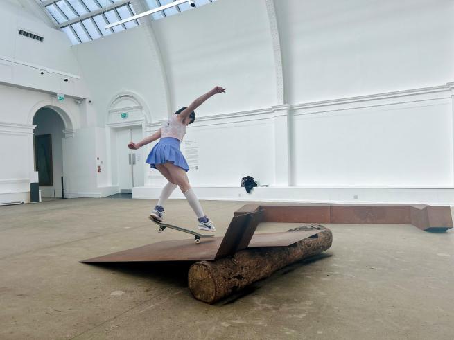 Image shows Ro Elliott skating on steel sculptures at Leeds Art Gallery. They are wearing a purple skirt that flares with the movement and arms outstretched