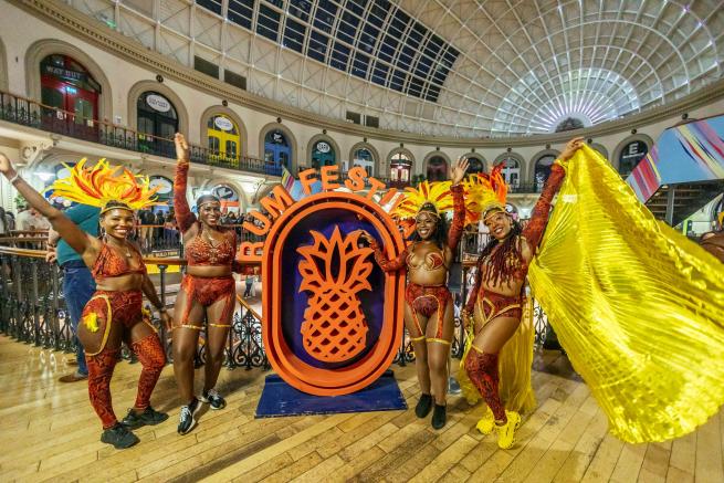 Four colourfully dressed carnival dancers surrounding large 'Rum Fest' wooden logo featuring a pineapple