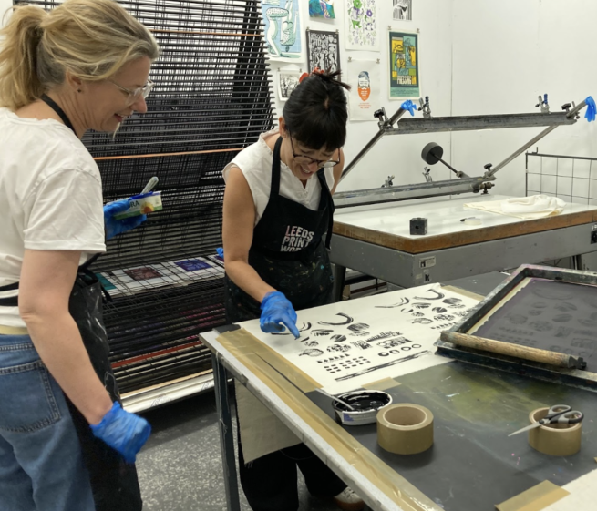 Two women looking at a piece of fabric screen printed with black abstract shapes, surrounded by screen printing equipment