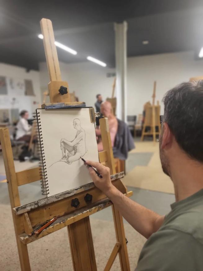 A point of view image of an artist sat at an easel drawing a life model