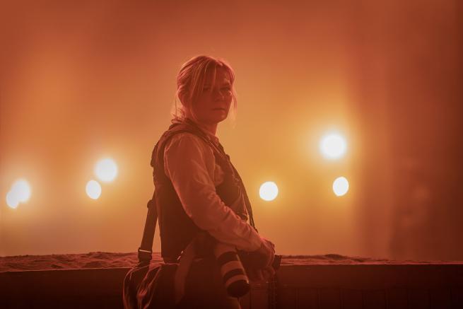 A woman in a bullet proof vest with a camera at her hip stands surrounded by lights and smoke