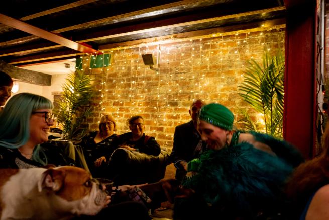 A burlesque performer with green feather fans interacts with a bulldog during a burlesque show. Smiling audience members in the background.