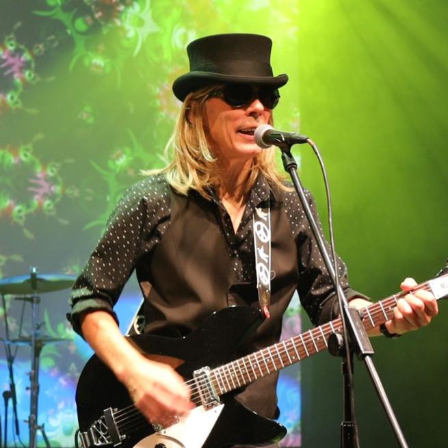 Image from Heartbreak: The Tom Petty Show of a man in a black top hat and shirt playing a black electric guitar, singing into a microphone on a stand.