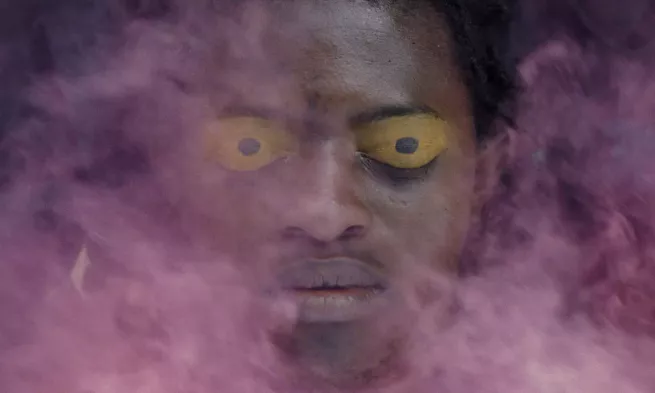 Close-up of a man's face amidst a cloud of pink smoke, his eyes are closed and hus eyeleids are painted yellow with a black spot in the middle.