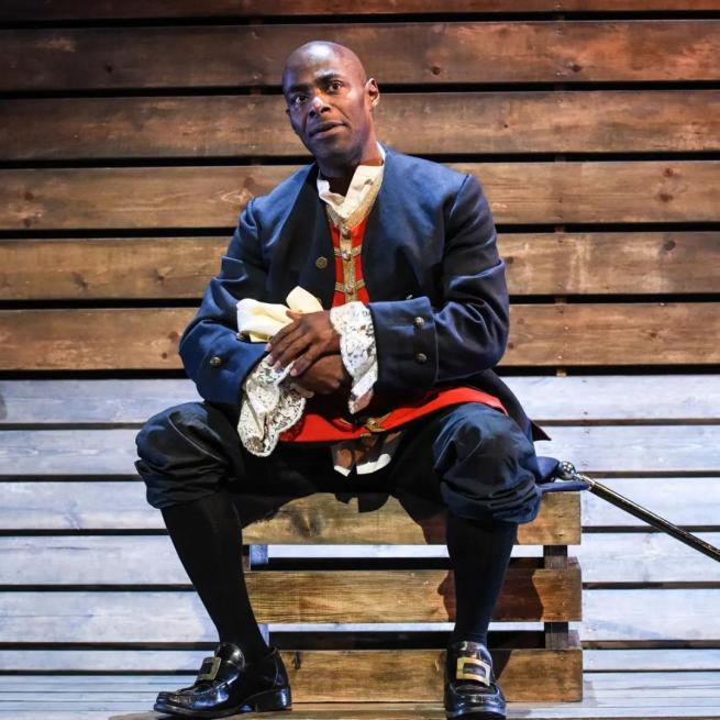 Paterson Joseph dressed in blue and red 18th Century military uniform sat on some wooden planks with a walking stick down to his left side.