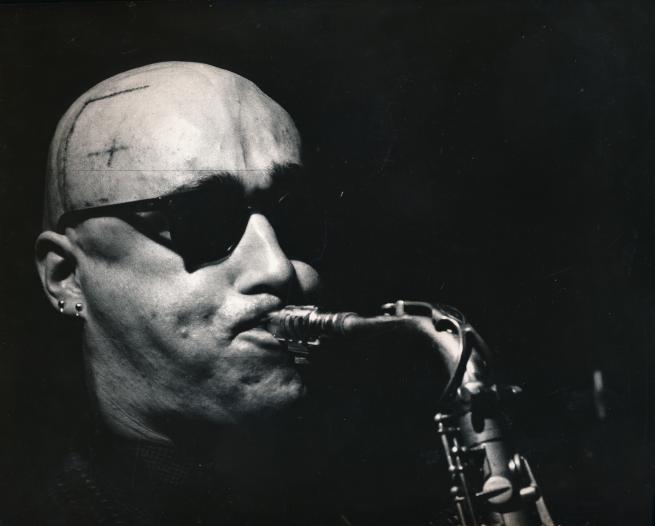 Xero Slingsby is picture in black and white, playing his saxophone. He has sunglasses on and lines drawn on his head from radiotherapy.