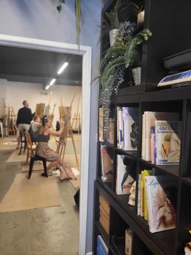 A corridor with a bookcase full of art books leading to an art studio with people sat drawing at their easels 