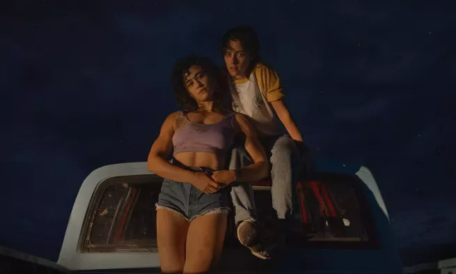 In a dark night, Jackie and Lou face the camera while leaning on a car.