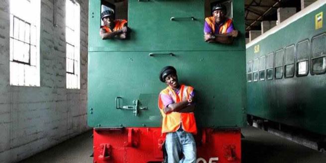 An image of the back of a green locomotive, with three men in orange vests. Two of the men are leaning out from the windows of the locomotive and one man is stood in front of it.