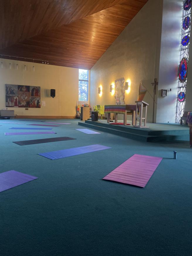 large room with different coloured yoga mats positioed in front of the alter in a semi circle. there is a stained glass window and another window leting light in and rising high to a slanted wooden ceiling.