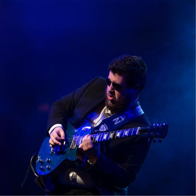 A person in sunglasses playing the guitar on stage as a misty blue light backlights the picture