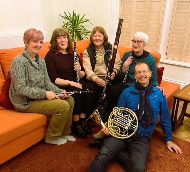 5 people seated and holding various musical instruments L to R clarinet, oboe, bassoon, flute, french horn