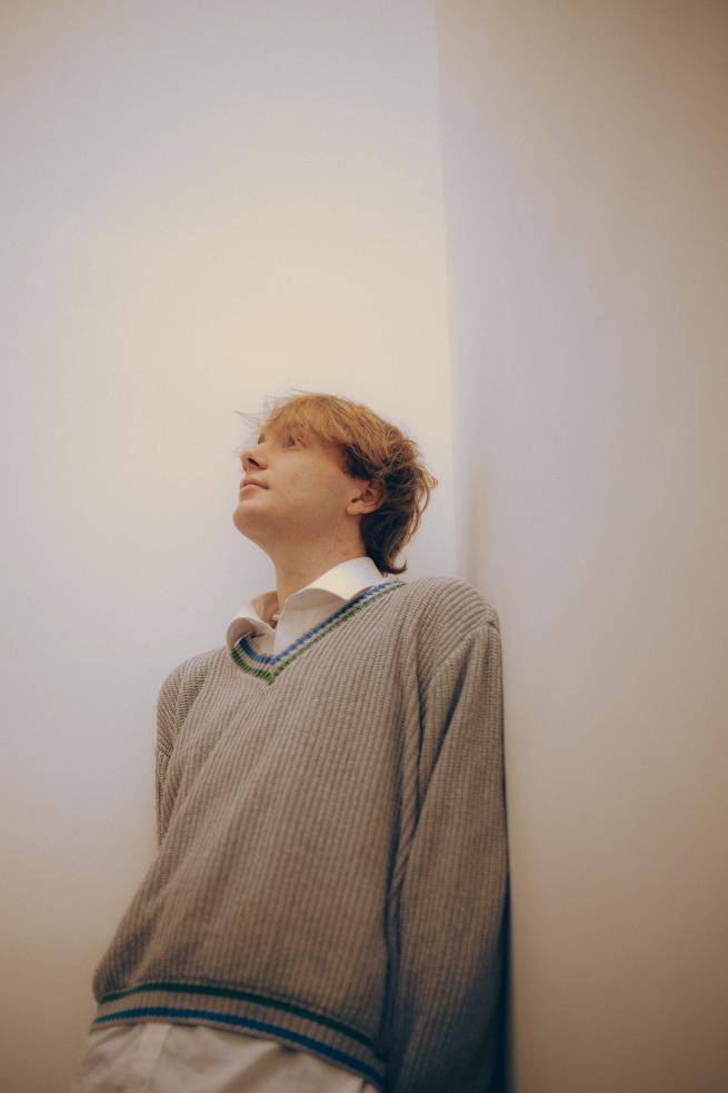 artist Jake Foster wearing a white shirt with a knit jumper overtop, leaning on a wall staring into the distance