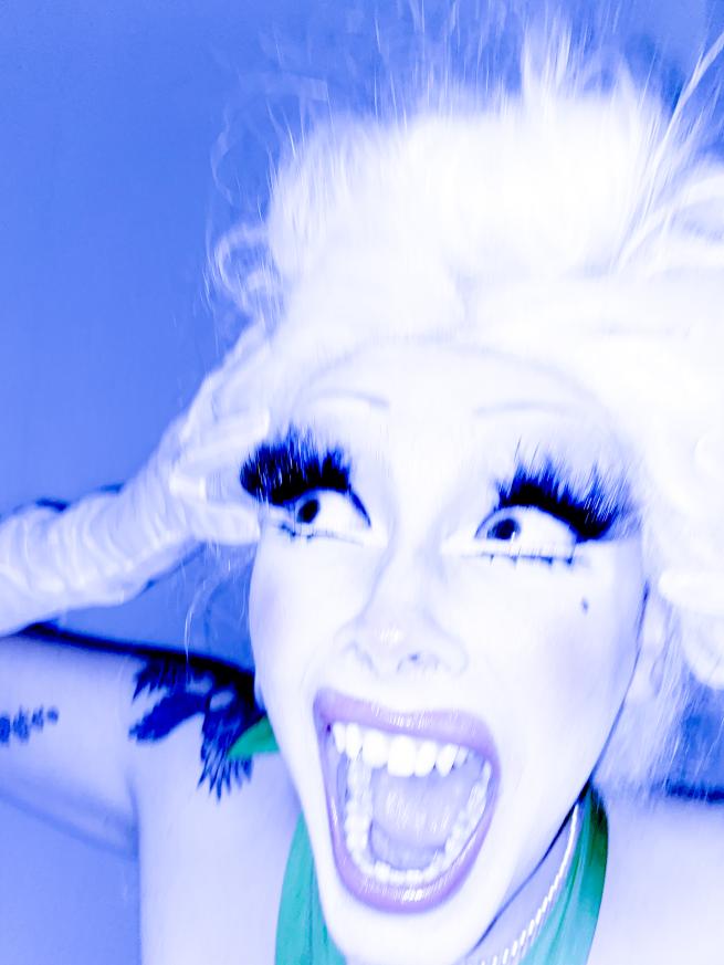An image of drag queen Liv Vicariously. They are wearing a platinum blonde wig styled into an up-do, making a shocked expression with both hands on their head. Their make up features overdrawn lips and eyeshadow, and gigantic lashes.