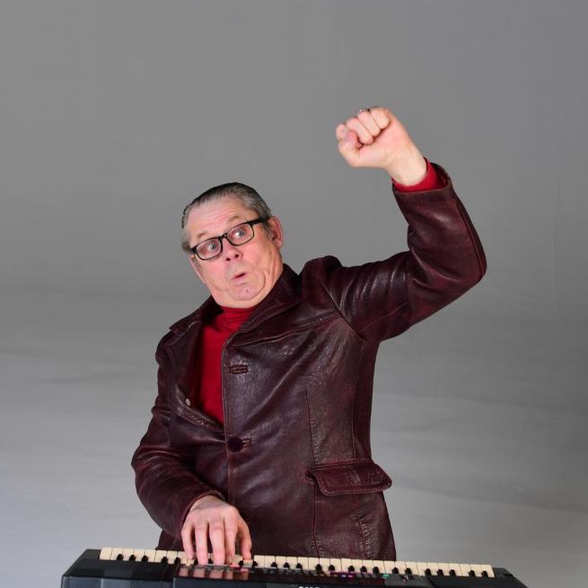 John Shuttleworth in a grey suit with his right hand on a keyboard and his left arm raised in the air in a fist.