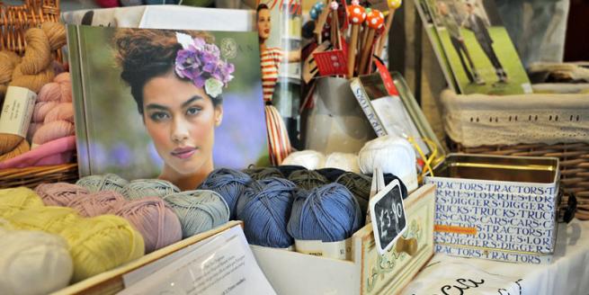 In the foreground of the image is balls of wool, on a table which has a pattern book and boxes of sale items.