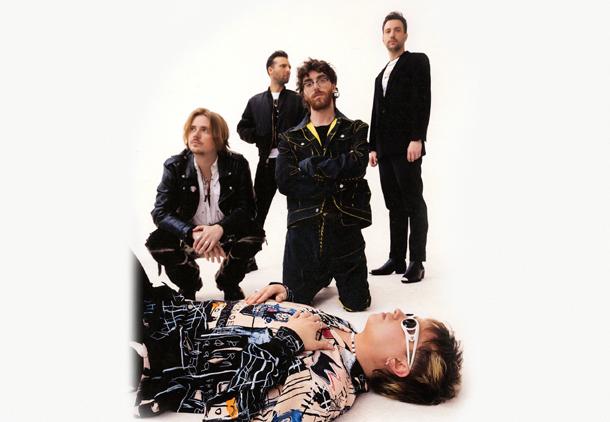 The five members of the band Nothing But Thieves are posing, with one lying on the ground and the other four standing or kneeling around him, all dressed in casual fashion.