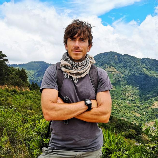 Simon Reeve standing in front of a view of some tropical-looking tree-covered mountains with his arms crossed.