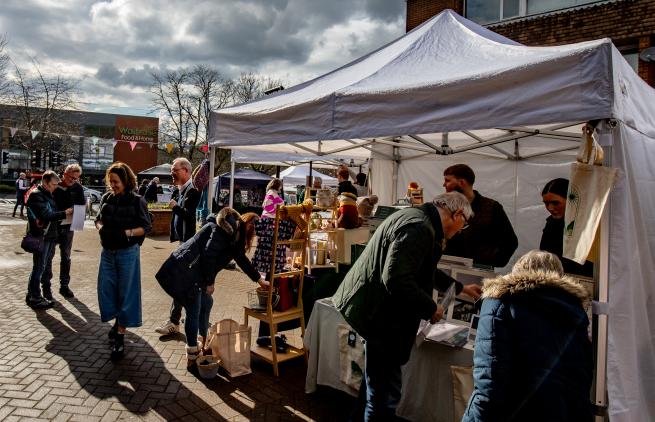 The photo shows an autumnal market day with people looking at a market stall and standing together chatting.